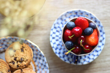 Plate of chocolate chip cookies, cup filled with strawberries, blueberries and cherries, open book and vase with gypsophila flowers on the table. Flat lay.