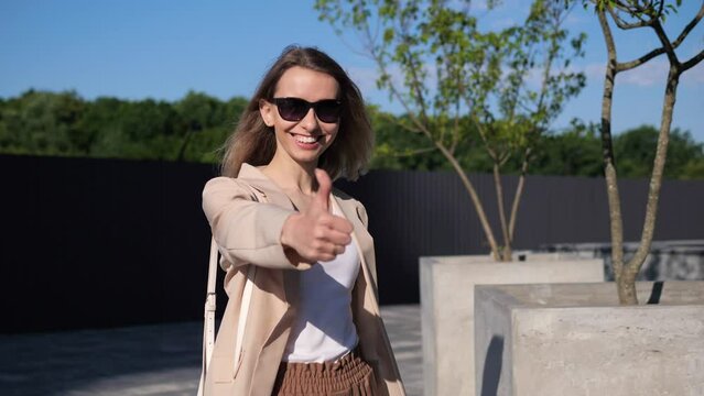 Positive ambitious young woman in sunglasses is showing thumbs up hand gesture standing outdoors with park area in background. Successful business people and entrepreneurship concept.