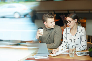 Thoughtful young business colleagues in smart casual clothes sitting at table in coffee shop and viewing papers while sharing ideas on strategy