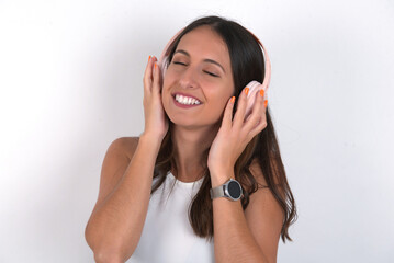 young beautiful caucasian woman wearing white Top over white background, smiles broadly feels very glad listens favourite music track via wireless headphones closes eyes.
