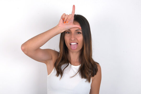 Funny young beautiful caucasian woman wearing white Top over white background, makes loser gesture mocking at someone sticks out tongue making grimace face.