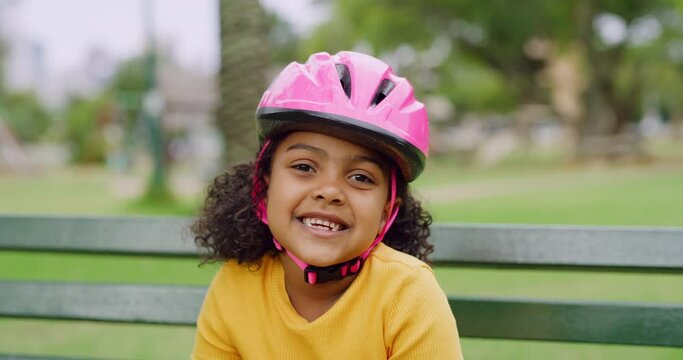 Portrait of happy Black girls face smiling and wearing a pink helmet and sitting on park bench outside. Cute positive child endorsing protective gear while having fun learning to ride bike or skate
