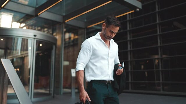 Mature businessman with neat beard wearing white shirt leaves the office in the financial district in the city. Successful man with mobile phone in his hands, after hard day's work