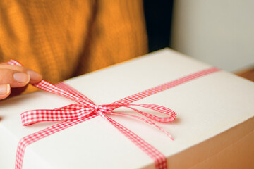Women's fingers hold red checkered ribbon on gift box. Festive surprise for Christmas, New Year or Birthday.