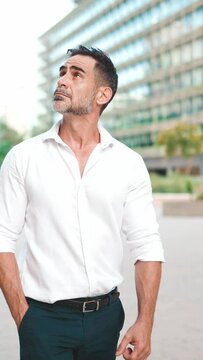 VERTICAL VIDEO: Mature businessman with neat beard wearing white shirt on his way to the office in the financial district in the city. Successful man Looks at the upper floors of modern buildings