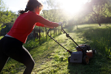 Mid Adult Woman in a Red Dress Uses a Motorized Lawn Mower on a Home Lawn at Sunset