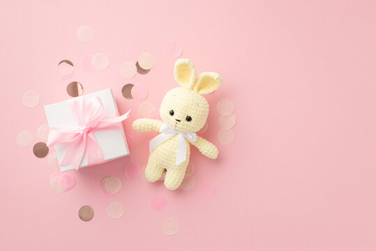 Baby concept. Top view photo of white giftbox with ribbon bow knitted bunny toy and shiny confetti on isolated pastel pink background