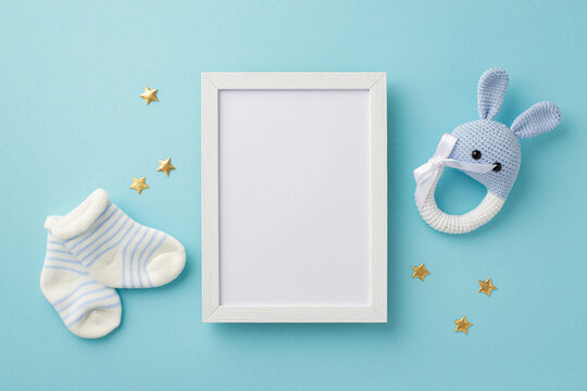Baby accessories concept. Top view photo of photo frame tiny socks knitted bunny rattle toy and gold stars on isolated pastel blue background with blank space