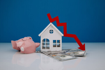 Wooden house and a red arrow down with broken piggy bank on money banknotes. The concept of...