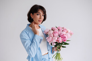 Beautiful brunette woman with short hair with bouquet of pions. Young girl with flowers. Portrait isolated. Pretty girl in blue suit smiling. Happy woman posing with flowers.