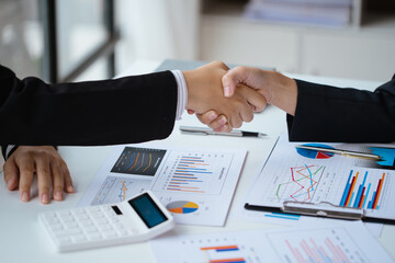 Close up of handshake above calculator paperwork that shows graft and chart of accounting and financial on table in office space.