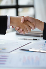 Close up of handshake above paperwork that shows graft and chart of accounting and financial on table in office space.