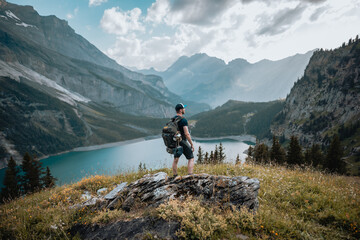 Un homme randonneur qui est en montagne lors d'une randonnée et qui regarde le magnifique panorama, avec un lac, des chemins et une forêt. - obrazy, fototapety, plakaty