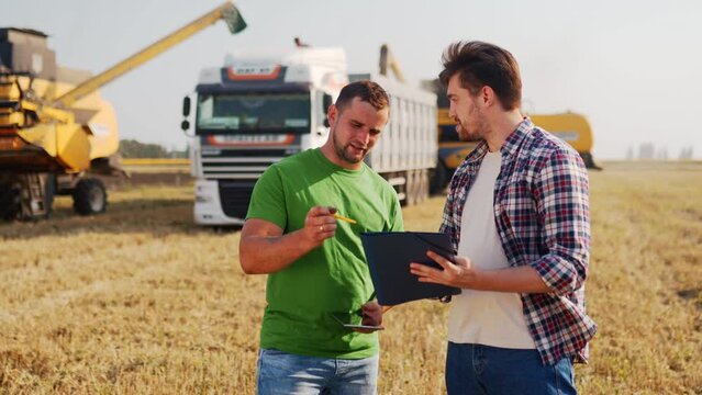 Farmer, logistics agent sign agreement on grain supply, shake hands in wheat field. Agronomist discuss agriculture business contract. Rancher, landlord negotiate with handshake. Harvesters load truck.