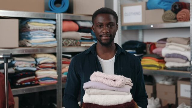 Portrait view of the multiracial male worker looking at the camera and stretching stack of clothes while working at the donating center or second hand. Humanitarian aid concept