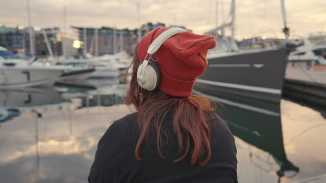 Unrecognizable young woman in headphones enjoying beautiful sunset over the sea, rear view. Slow motion