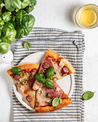 Three pieces of pizza in white plate on striped napkin, glass , salt shaker and basil leaves on white table. Top view