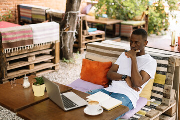 Fototapeta na wymiar Serious, concentrated and enthusiastic african afro american businessman sitting on bench with pillows using laptop and drinking coffee in street internet cafe. Male noting and calculating data online