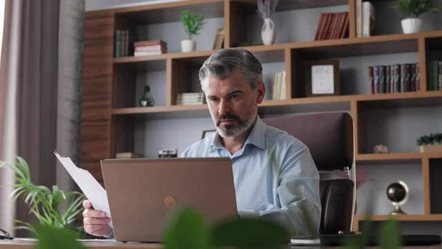 Portrait Of Adult Gray-haired Bearded Businessman Working With Laptop At Home Office. Texting Messages, Doing Paperwork, Calculating Monthly Expenses. Business Concept.