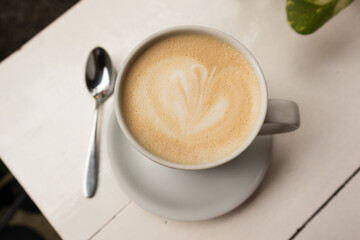 cup of coffee with milk with a drawing, in the background a white table, with a spoon and leaves of a plant