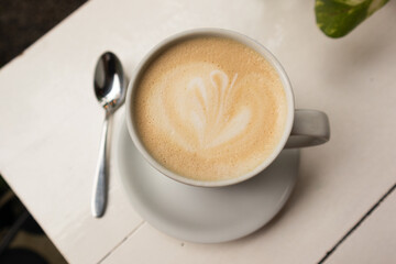 cup of coffee with milk with a drawing, in the background a white table, with a spoon and leaves of...
