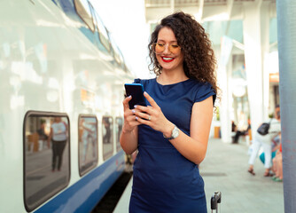 Attractive cute curly hair girl in blue dress and trendy glasses using phone on train station 