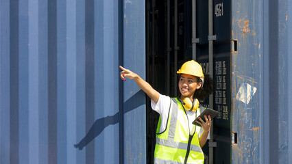 Young African American female trainee uses tablet to check cargo containers and stocks in a logistic warehouse. Female worker in hardhat, earmuff, and safety suit stands next to a blue container.