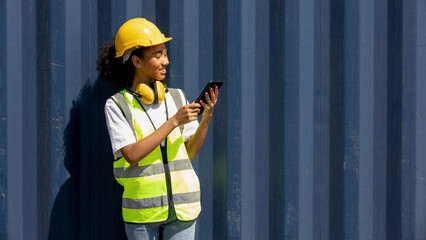 Young African American female trainee uses tablet to check cargo containers and stocks in a logistic warehouse. Female worker in hardhat, earmuff, and safety suit stands next to a blue container.