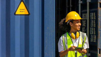 Young African American female trainee check cargo containers and stocks in a logistic warehouse. Female worker in hardhat and safety suit stands next to a blue container. Factory driver with earmuff - obrazy, fototapety, plakaty