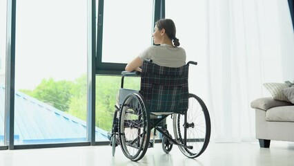Smiling young woman in wheelchair relaxing at home near window and looks outside. Leisure at home
