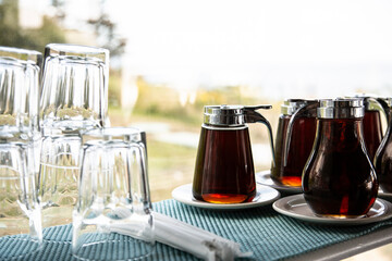 Amber maple syrup in glass serving dispensers on saucers next to glassware by a window.