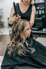 Fototapeta premium hands of a hairdresser with a curling iron make a hairstyle to a curly girl in a professional beauty salon