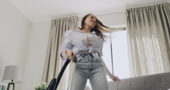 One young woman from below dancing with joy and cleaning a carpet with vacuum cleaner in a lounge at home. Cheerful and energetic woman having fun while doing chores and housework for tidy apartment