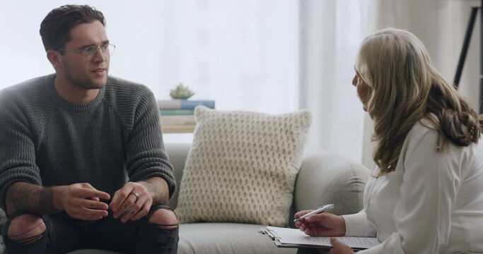 Young man on a couch in a session with a psychologist. Talking to his therapist about his problems and working through difficult emotions or memories while she makes notes
