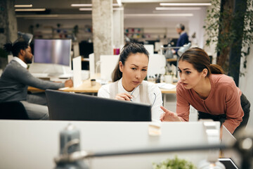 Two businesswomen talk while reading e-mail on computer in office.