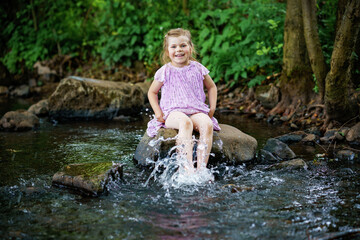 Cute little toddler girl having fun by a river on warm and sunny summer day. Happy excited preschool child splashing with water in forest stream creek.
