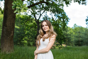 Naklejka na ściany i meble Beautiful young Russian girl dressed in a white dress, walks through the forest among the oaks in the rays of the sunset