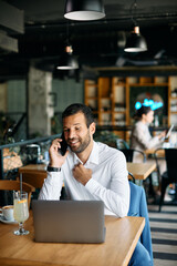 Happy entrepreneur talking on the phone while using laptop in cafe.