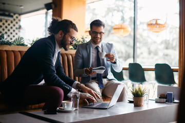 Happy entrepreneur using laptop while drinking coffee with coworker in cafe.