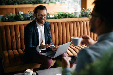 Young happy entrepreneur works on laptop while talking to coworker during business meeting in cafe.
