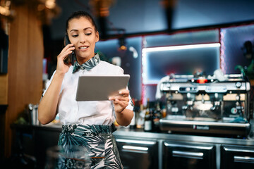 Young waitress uses touchpad while communicating on cell phone in pub.