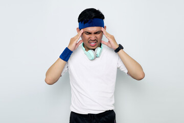 Stressed young Asian sportman in blue headband and sportswear white t-shirt with headphones,touching his temples and having headache isolated on white background. Workout sport concept