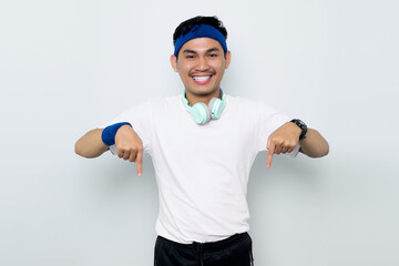 Smiling young Asian sportman in blue headband and white t-shirt with headphones, pointing fingers...
