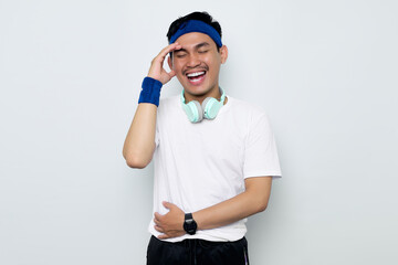young Asian sportman in blue headband and sportswear white t-shirt with headphones, laughing holding his hand on stomach can't stop laughing after hearing funny anecdotes isolated on white background