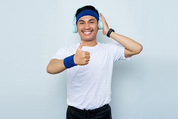 Portrait of cheerful young Asian sportman in blue headband and sportswear white t-shirt while listening music favorite with headphones, showing thumb up gesture isolated on white background