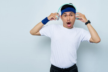 Shocked young Asian sportman in blue headband and sportswear white t-shirt while listening music favorite with headphones keeping mouth open isolated on white background. Workout sport concept