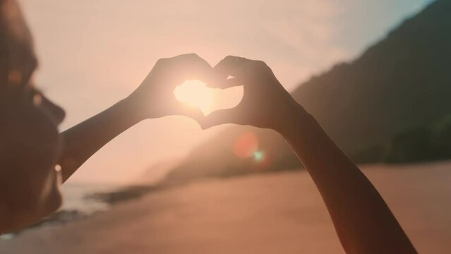 Young happy Caucasian woman teen with hair blowing in wind smiling making heart symbol with hands demonstrating love for this vacation spot hoping to return in future stands on beach near sea