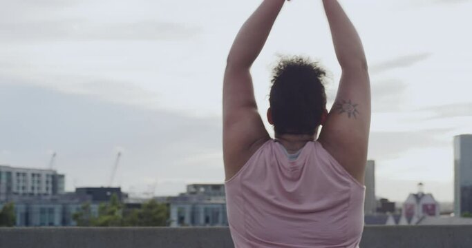 One plus size woman with arms up cheering after her early morning jog. Young curvy female celebrating her fitness milestone. Happy girl in active wear standing with her back, overlooking the city