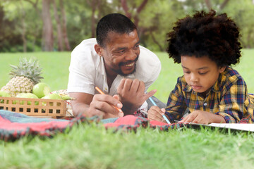 Happy smiling African American father and son go out for picnic outdoor in park on holiday, boy with curly hair drawing picture, lying on green grass with dad, joyful family playing outside in nature.