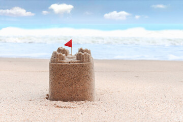 Fototapeta na wymiar Sand castle with red flag on tropical sandy summer ocean beach with beautiful blue sky as background, relaxing outdoor vacation on sea beach island.